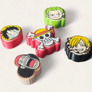 Puede incluir: Colección de gomas de borrar coloridas con forma de personajes de la serie de anime One Piece. Las gomas muestran varios personajes con expresiones faciales distintas y colores vibrantes, incluyendo rojo, amarillo, verde y negro.