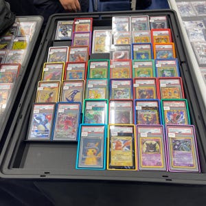 May include: A display case filled with graded trading cards, each encased in a protective holder with a coloured border. The cards feature various characters and designs, with a focus on vibrant colours and detailed artwork. The case is black and sits on a dark surface.