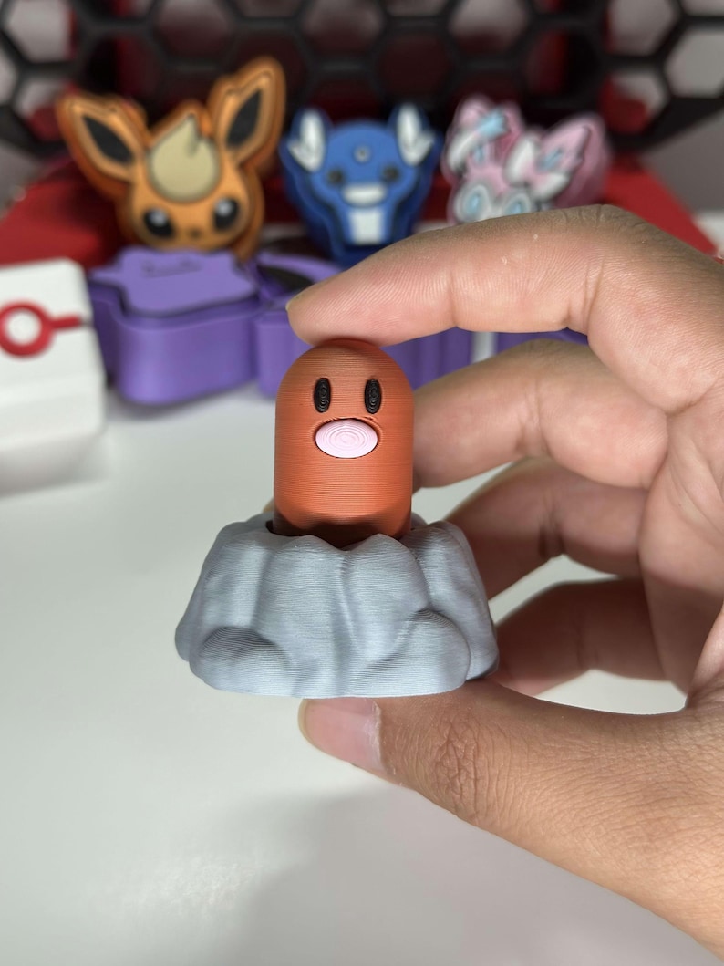 May include: A 3D-printed Diglett figure, a character from the Pok&eacute;mon franchise, in a light orange color with a pink mouth and black eyes, emerging from a gray rock formation. Other Pok&eacute;mon figures are visible in the background.