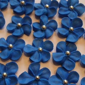 May include: A collection of blue sugar flowers with silver centers. The flowers are arranged in a random pattern on a white surface.