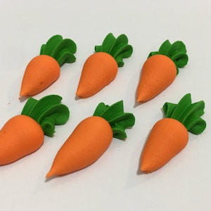 May include: Six orange carrot-shaped decorations with green leafy tops are arranged on a white surface. The carrots are a vibrant orange color, and the green leaves are a contrasting shade. These decorative carrots are likely for baking or crafting.
