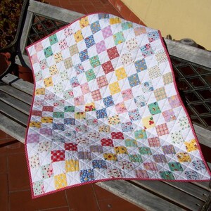 Op de afbeelding: Een patchworkdeken met een witte achtergrond en vierkanten in verschillende kleuren en patronen, waaronder rood, geel, blauw en groen. De deken is afgewerkt met een rode en witte rand.