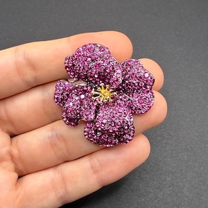 May include: A large, flower-shaped brooch covered in small, sparkling magenta crystals. The center of the flower features a gold-colored, star-like design. The brooch is held in a hand against a dark background, showcasing its intricate details.