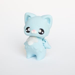 May include: A light blue cat figurine with large black eyes, a pink nose, and small wings. The cat has a fluffy white chest and is standing upright. The figurine is made of a smooth material and is set against a white background.