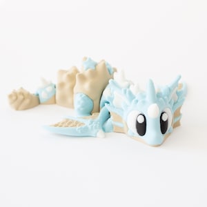 May include: A light blue and tan dragon figurine with large black eyes and white accents. The dragon has a stylized design with a long body and a detailed wing. The figurine is lying down.