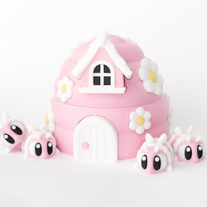 May include: A pink, whimsical house-shaped container with a white door, window, and roof. The house is decorated with white and yellow flower accents. Several pink, cartoonish bug-like figures with large eyes surround the house.