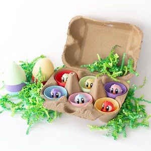 May include: A brown cardboard egg box containing six toy birds in small, brightly coloured bowls. Two decorative eggs and green paper grass are on a white surface. The birds are in red, blue, pink, yellow, green, and purple bowls.