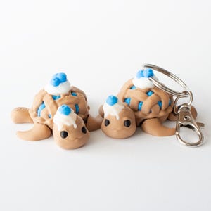 May include: Two whimsical turtle keychains. The turtles are brown with blue and white accents, resembling a pie crust and whipped cream. One keychain has a silver clasp.