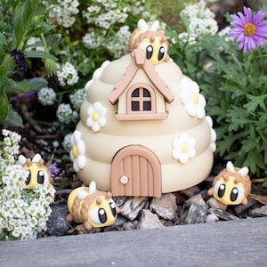May include: A miniature beehive with a small house on top, surrounded by white flowers and four small bee figurines. The beehive is painted in a light brown color with white accents.