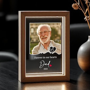 May include: A wooden framed photo with a warm LED light. The photo features a smiling man with glasses. The text on the frame reads "Forever in our hearts Dad 2023" with a heart symbol. A vase of dried flowers sits to the right.