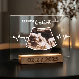 May include: A clear acrylic frame with a wooden base, displaying a black and white ultrasound image. The frame has a white inscription that reads "At First Heartbeat" with a heart symbol. The date "02.27.2025" is engraved on the wooden base.