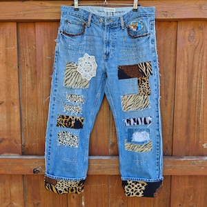 May include: Light blue denim jeans with various animal print patches and a white lace doily. The jeans have rolled-up cuffs with a leopard print design and brown leather accents on the pockets.