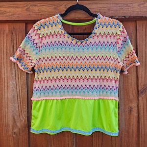 May include: A colorful, short-sleeved top with a V-neck design. The top features a vibrant zigzag pattern in various colors, including pink, orange, blue, and green. The lower portion is a bright green fabric, and the edges are trimmed with pink and blue detailing.