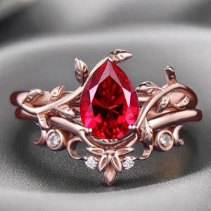 May include: A rose gold ring featuring a pear-shaped, vibrant red gemstone. The ring's design incorporates delicate leaf and vine motifs, with small, clear accent stones adding sparkle. The ring is set against a soft gray background.