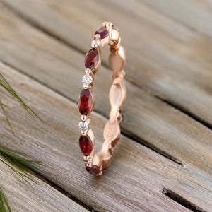 May include: A rose gold band ring featuring an alternating pattern of marquise-cut deep red garnet gemstones and small round-cut clear white diamonds. The metal band has a polished finish and a delicate, slender profile with a continuous design.