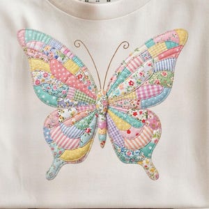 May include: A cream-coloured t-shirt with a large, colourful butterfly design. The butterfly is made up of various fabric patterns in pastel shades of pink, blue, yellow, and green. The shirt is neatly folded on a wooden surface.