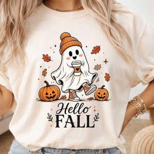May include: A cream-colored t-shirt featuring a graphic of a white ghost wearing an orange beanie and sneakers while holding a coffee cup. The design includes two jack-o'-lanterns, falling autumn leaves, and the text Hello FALL in black.