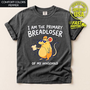 May include: A charcoal gray short-sleeve t-shirt featuring a graphic of a yellow mouse wearing sunglasses and holding a piece of bread. The text reads I AM THE PRIMARY BREADLOSER OF MY HOUSEHOLD in white capital letters above and below the mouse.