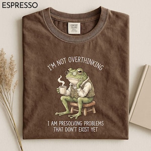 May include: A brown crewneck t-shirt featuring a graphic of a green frog wearing a vest and glasses, sitting on a stool while holding a coffee cup. The text reads I'M NOT OVERTHINKING I AM PRESOLVING PROBLEMS THAT DON'T EXIST YET. Comfort Colors label.