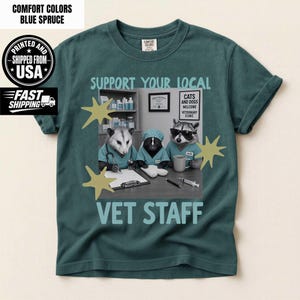 May include: A blue spruce t-shirt featuring a graphic of three animals dressed as veterinary staff. The text reads SUPPORT YOUR LOCAL VET STAFF, CATS AND DOGS WELCOME VETERINARY CLINIC. The animals include an opossum, a skunk, and a raccoon.