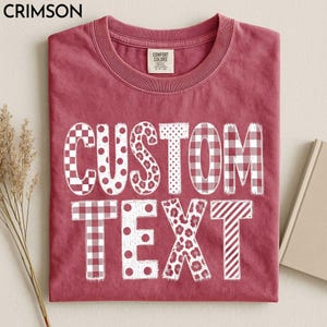 May include: A crimson red crew neck t-shirt featuring the white text CUSTOM TEXT in a bold, blocky font. Each letter displays a unique pattern, including checkered, polka dot, leopard print, and striped designs. A Comfort Colors label is visible.