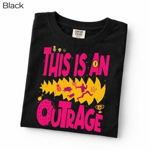 May include: A black t-shirt featuring a graphic print with the text "THIS IS AN OUTRAGE" in large pink letters. The design includes a yellow jagged shape, a donut, a trophy, a pineapple, skulls, and a silhouette of a person running with a torch.