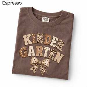 May include: An espresso-coloured t-shirt with a crew neckline and a Comfort Colors label. The front features the text KINDERGARTEN in large, blocky letters with alternating leopard print and polka dot patterns, accompanied by a matching leopard print bow.