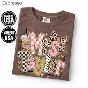 Peut inclure: Un t-shirt marron avec un motif graphique portant le texte Mrs Taylor. Le design comprend une tasse à café, un nœud à imprimé léopard et divers motifs incluant des carreaux roses et blancs, des carreaux noirs et blancs, des pois et des paillettes dorées.