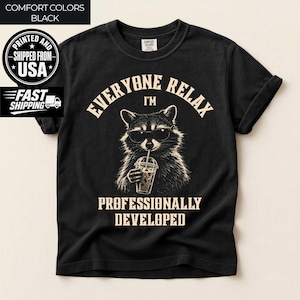 May include: A black t-shirt featuring a graphic of a raccoon wearing sunglasses and drinking from a plastic cup with a straw. The text reads "EVERYONE RELAX I'M PROFESSIONALLY DEVELOPED" in a beige, arched, and block-style font.