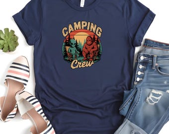 Puede incluir: Camiseta azul marino con un gráfico de un extraterrestre y Bigfoot sentados en sillas de camping, con las palabras "Camping Crew". El diseño está sobre un fondo de puesta de sol.