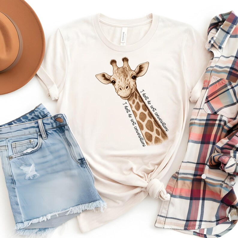 May include: A cream-coloured t-shirt with a giraffe illustration and the text "I don't do soft conversations." The giraffe is in brown and tan shades. The shirt is styled with denim shorts, a brown hat, and a plaid shirt.