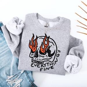 Może przedstawiać: Jasnoszara bluza z nadrukiem czaszki z pomarańczowymi płomieniami i napisem "Everything's Fine". Srebrny naszyjnik jest zarzucony na bluzę, która leży obok pary niebieskich dżinsów.