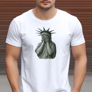 Op de afbeelding: Wit T-shirt met een grijze afbeelding van het Vrijheidsbeeld dat haar ogen bedekt. De afbeelding heeft een distressed design. Het T-shirt heeft een ronde hals en korte mouwen.