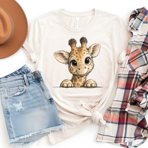 May include: A cream-coloured short-sleeve t-shirt featuring a central graphic of a cartoon giraffe with large brown eyes and a spotted pattern. The shirt is displayed alongside a brown hat, light blue denim shorts, and a plaid button-down shirt.
