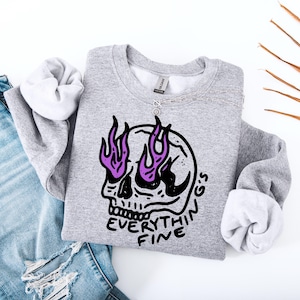Może przedstawiać: Jasnoszara bluza z czarną grafiką czaszki i fioletowymi płomieniami. Tekst "EVERYTHING'S FINE" jest wydrukowany pod czaszką. Srebrny naszyjnik jest zarzucony na bluzę.