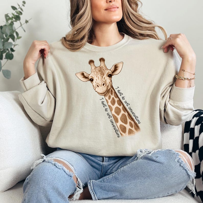 May include: A beige sweatshirt with a giraffe graphic and the text "I don't do soft conversations." The giraffe is depicted with a brown and tan spotted pattern. The sweatshirt is a neutral colour, suitable for casual wear.