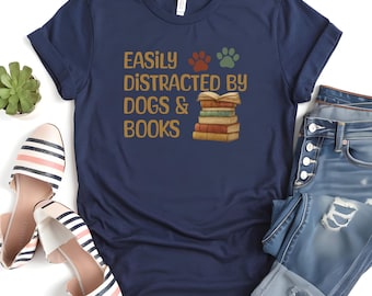 May include: Navy blue t-shirt with the text "EASILY DISTRACTED BY DOGS & BOOKS" in gold lettering. The design includes paw prints and a stack of books. The shirt is paired with blue jeans and striped shoes.