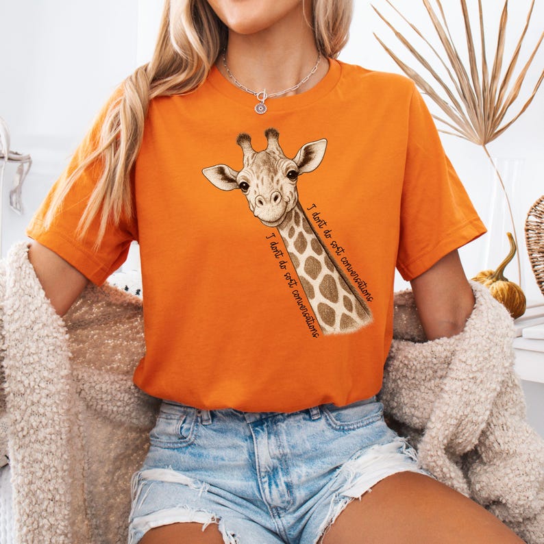 May include: An orange t-shirt with a giraffe graphic and the words "I don't do soft conversations." The giraffe has a brown and white spotted pattern. The shirt is a short-sleeved, casual style.