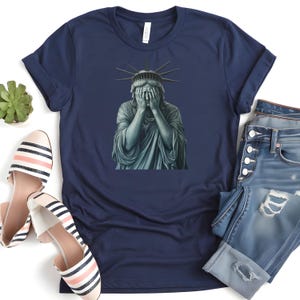 May include: A navy blue t-shirt featuring a graphic of the Statue of Liberty with her hands over her face.