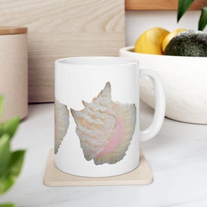 Pode incluir: Caneca de cerâmica branca com um design de três conchas. As conchas são pintadas em tons de rosa, branco e bege.