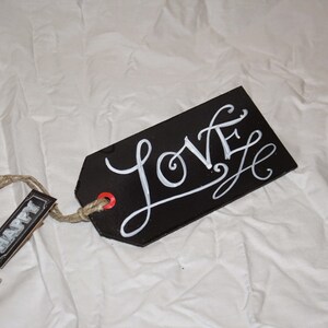 May include: Black tag with a white handwritten word "LOVE" on it. The tag is attached to a twine string with a smaller tag that says "HAPPY" on it.