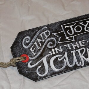 May include: Black chalkboard tag with white lettering that reads "Find Joy in the Journey". The tag is attached to a piece of twine.