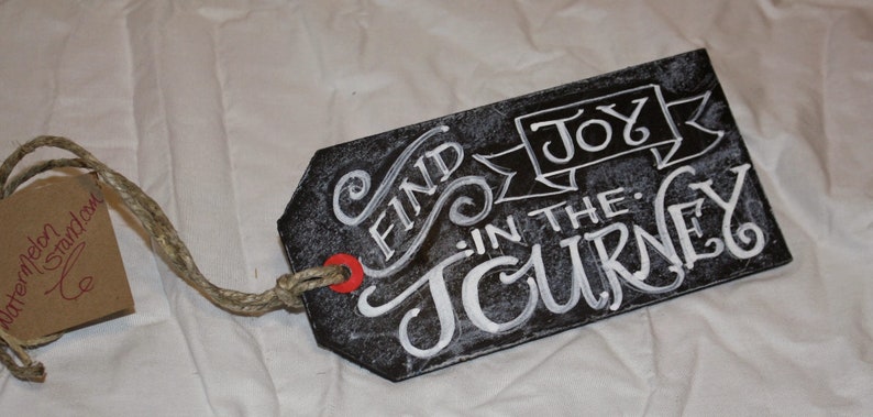 May include: Black chalkboard tag with white lettering that reads "Find Joy in the Journey". The tag is attached to a piece of twine.