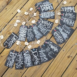May include: A collection of 20 black chalkboard tags with white handwritten words on them. The words include "Believe", "Dream", "Faith", "Love", "Strength", "Hope", "Courage", "Create", "Destiny", "Trust", "Smile", "Guest Room", "Vintage", "Breathe", "Family", "True", "Heart Mind", "I Write", "Farm Eggs", and "Desire".