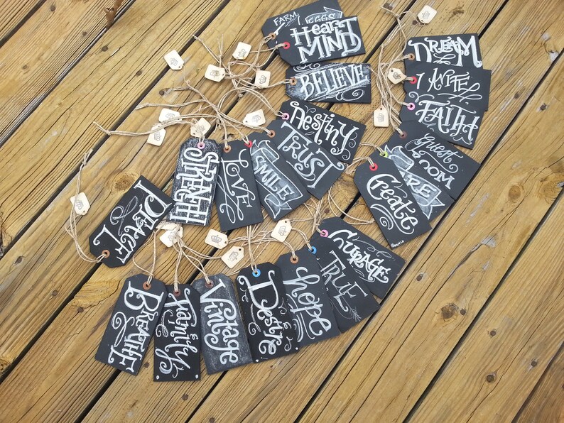May include: A collection of 20 black chalkboard tags with white handwritten words on them. The words include "Believe", "Dream", "Faith", "Love", "Strength", "Hope", "Courage", "Create", "Destiny", "Trust", "Smile", "Guest Room", "Vintage", "Breathe", "Family", "True", "Heart Mind", "I Write", "Farm Eggs", and "Desire".