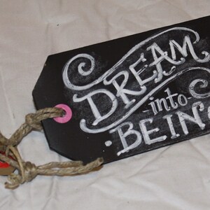 May include: A black chalkboard tag with a white rope attached. The tag has white chalk lettering that reads "Dream into Being."