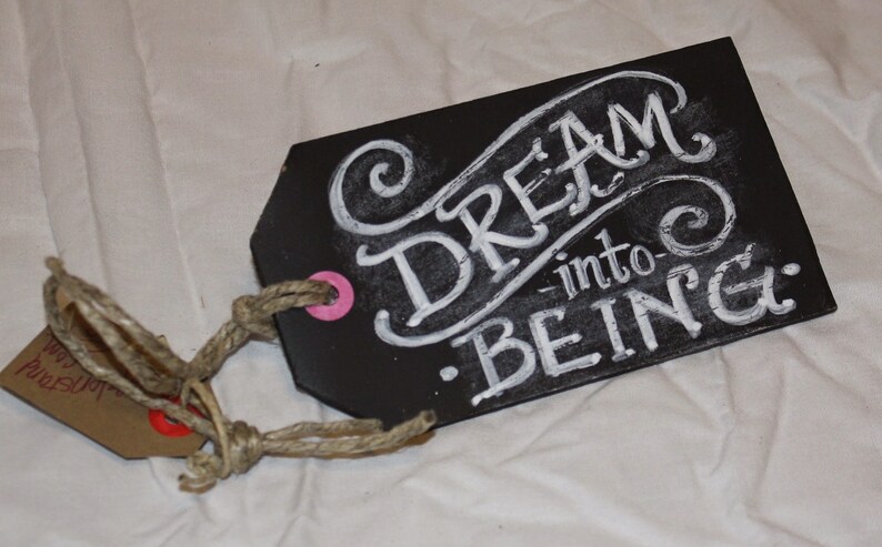 May include: A black chalkboard tag with a white rope attached. The tag has white chalk lettering that reads "Dream into Being."