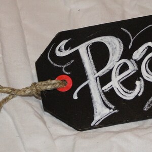 May include: Black chalkboard tag with white chalk lettering that says "Peace". The tag is tied with twine and has a red dot on the top.