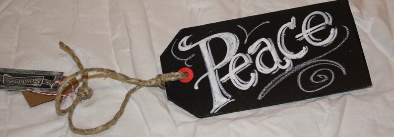 May include: Black chalkboard tag with white chalk lettering that says "Peace". The tag is tied with twine and has a red dot on the top.