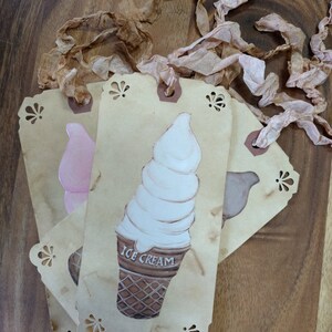May include: Four brown paper gift tags with scalloped edges. Each tag has a different image of a dessert: a pink ice cream cone, a white ice cream cone with the text "ICE CREAM", a brown ice cream cone, and a brown bird. The tags are tied together with brown and pink ribbon.